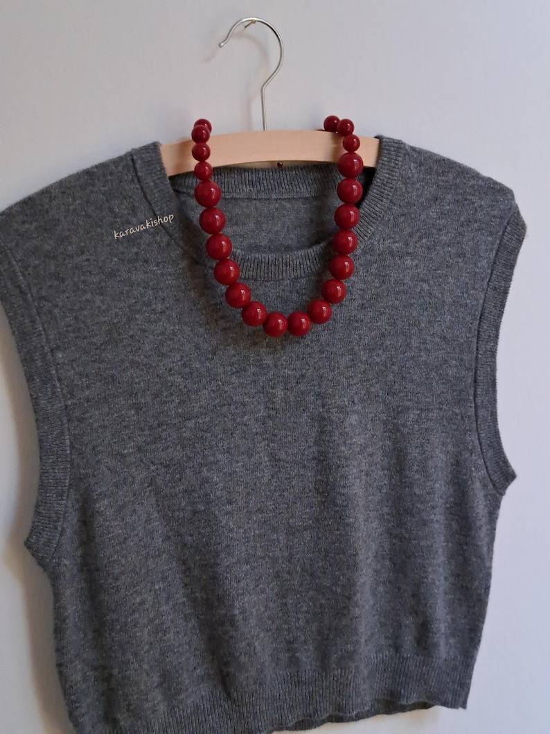 Pu&ograve; includere: Un gilet in maglia grigio senza maniche con scollo rotondo, caratterizzato da una collana di grandi perle rosse rotonde e lucide drappeggiata sul colletto. Il testo karavakishop &egrave; stampato in piccole lettere bianche sul lato superiore sinistro.