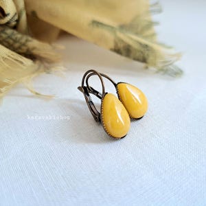May include: A pair of teardrop-shaped earrings featuring smooth, opaque yellow stones set in antique bronze-toned metal frames with lever-back closures. The text karavakishop is visible on the left side of the earrings, which are displayed together.