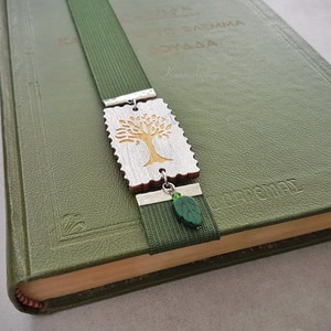May include: A green bookmark with a silver metal charm featuring a wooden tree design. The charm is attached to the bookmark with a silver metal ring. A green glass leaf charm hangs from the bottom of the bookmark.