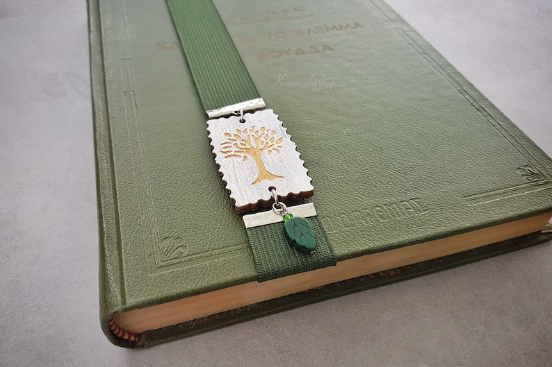 May include: A green bookmark with a silver metal charm featuring a wooden tree design. The charm is attached to the bookmark with a silver metal ring. A green glass leaf charm hangs from the bottom of the bookmark.