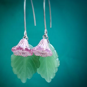May include: A pair of dangle earrings featuring silver-toned curved ear wires. Each earring displays a small, translucent pink bell-shaped flower bead suspended above a larger, semi-translucent light green leaf-shaped bead with textured veins.