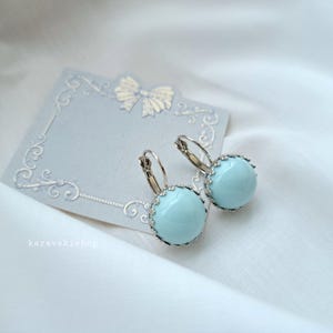 May include: A pair of light blue cabochon earrings with silver-toned scalloped settings and lever-back closures, displayed on a light gray card featuring a decorative bow and scrollwork border with the text karavakishop printed in white.