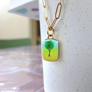 May include: A gold-toned paperclip chain necklace featuring a rectangular pendant with rounded corners. The pendant displays a painted scene of a single black tree silhouette against a bright green and yellow background, suspended by a small gold ring.