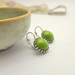 Op de afbeelding: Een paar oorbellen met klapbrisure, voorzien van gladde, ronde, limoengroene cabochons in decoratieve, zilverkleurige metalen zettingen met een geschulpte rand, geplaatst naast een kleine, lichtgroene keramische kom met een gele rand.