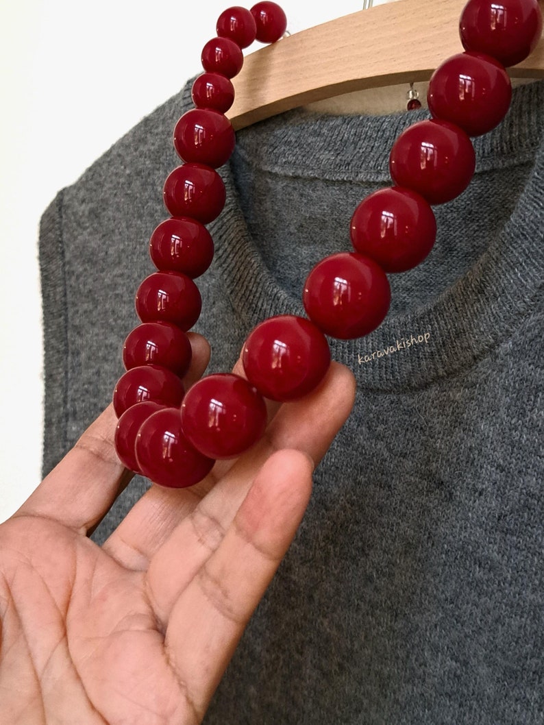 Pu&ograve; includere: Una collana composta da grandi perle sferiche rosso scuro, lucide e lisce. Le perle hanno una superficie riflettente. Il testo karavakishop &egrave; visibile in piccole lettere bianche vicino al centro della collana sul tessuto grigio.