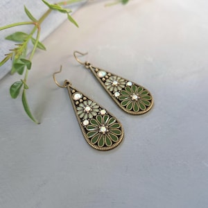 Pode incluir: Um par de brincos pendentes em forma de gota com uma armação de metal cor de bronze. Cada brinco apresenta dois motivos florais preenchidos com esmalte verde-oliva e sálvia, acentuados por pequenos strass claros e iridescentes no centro.