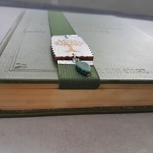 May include: A green book with a bookmark featuring a wooden tree charm and a green leaf charm. The bookmark is attached to a green elastic band.