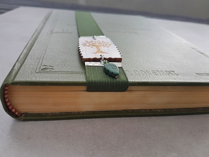 May include: A green book with a bookmark featuring a wooden tree charm and a green leaf charm. The bookmark is attached to a green elastic band.