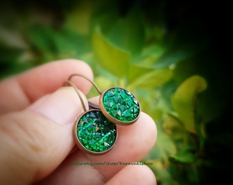 May include: A pair of round earrings featuring faceted emerald green crystals set in antique bronze metal frames with curved ear wires. The circular stones have a textured, sparkling surface that reflects light, held by a metallic bezel setting.