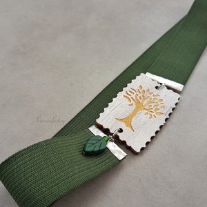May include: A green elastic bookmark with a wooden charm featuring a gold tree design. The charm is attached to the bookmark with a silver metal connector and a green leaf bead.