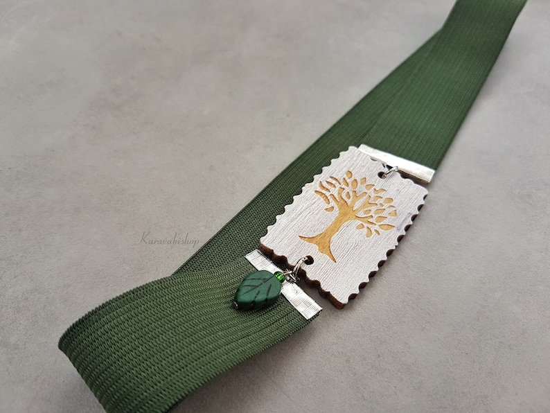 May include: A green elastic bookmark with a wooden charm featuring a gold tree design. The charm is attached to the bookmark with a silver metal connector and a green leaf bead.