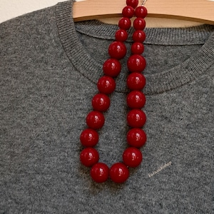 Pu&ograve; includere: Una collana composta da due fili di perle sferiche rosso scuro lucide di varie dimensioni, disposte in un motivo a gradazione. Le perle sono infilate insieme e poggiano su un maglione in maglia grigio m&eacute;lange testurizzato.