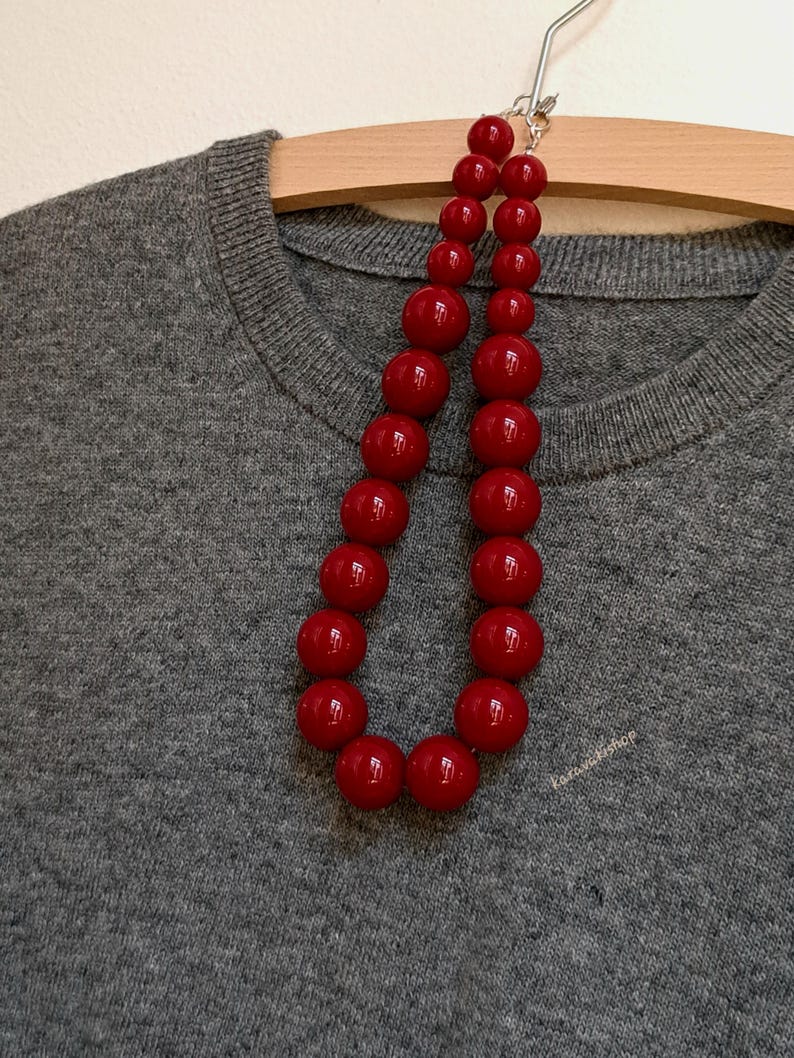 Pu&ograve; includere: Una collana composta da due fili di perle sferiche rosso scuro lucide di varie dimensioni, disposte in un motivo a gradazione. Le perle sono infilate insieme e poggiano su un maglione in maglia grigio m&eacute;lange testurizzato.