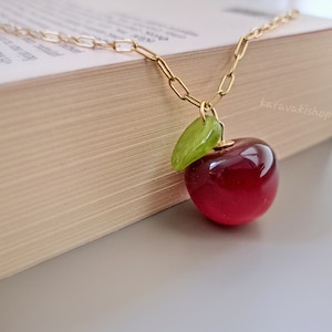 Può includere: Una collana a catena color oro con un ciondolo a forma di ciliegia rossa lucida e traslucida, con una piccola foglia verde brillante attaccata. Il ciondolo pende da una sottile catena a maglie metalliche e il testo karavakishop è visibile sullo sfondo.