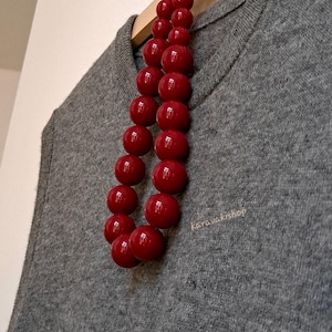 Pu&ograve; includere: Una collana composta da due fili di grandi perle sferiche rosso scuro e lucide. La collana &egrave; appoggiata su un maglione di maglia grigio. Il testo "karavakishop" &egrave; stampato in piccole lettere chiare sul tessuto del maglione.