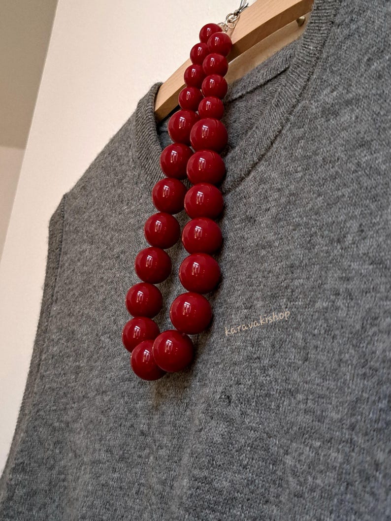 Pu&ograve; includere: Una collana composta da due fili di grandi perle sferiche rosso scuro e lucide. La collana &egrave; appoggiata su un maglione di maglia grigio. Il testo "karavakishop" &egrave; stampato in piccole lettere chiare sul tessuto del maglione.