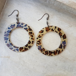 Könnte beinhalten: Ein Paar große, runde Creolen mit Leopardenmuster. Die Ohrringe sind aus klarem Kunststoff mit einem braunen und schwarzen Leopardenmuster. Die Ohrringe haben silberne Haken.