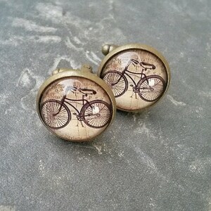 Może przedstawiać: Para okrągłych spinek do mankietów z ilustracją roweru w stylu vintage pod przezroczystą szklaną kopułą. Spinki mają metalową ramkę w kolorze brązu oraz tło przypominające postarzany papier w odcieniu sepii z niewyraźnym tekstem.