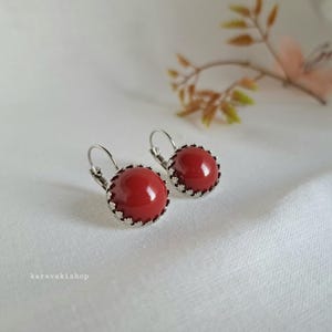 Op de afbeelding: Een paar oorbellen met een klapmechanisme, voorzien van gladde, ronde, dieprode cabochons in een geschulpte, zilverkleurige metalen zetting. De tekst karavakishop is zichtbaar in de linkerbenedenhoek van de afbeelding.