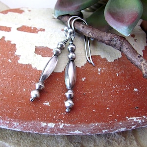May include: Silver dangle earrings with a combination of small round beads and elongated oval beads. The earrings hang from simple silver hooks. The design is simple and elegant, suitable for everyday wear.