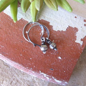 May include: A pair of silver hoop earrings with small, round, silver beads hanging from them. The earrings are on a terracotta-colored surface.