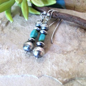 May include: Dangle earrings featuring a vertical arrangement of polished silver beads and cylindrical turquoise stones. The earrings are suspended from silver ear wires, showcasing a combination of metallic spheres and natural blue-green stone accents.
