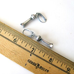 May include: A pair of silver earrings with a round bead and a long, thin, cylindrical pendant. The earrings are measured against a wooden ruler.