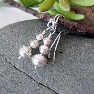 May include: Silver dangle earrings with three round beads on each earring. The beads are stacked on top of each other and have a slightly textured surface.