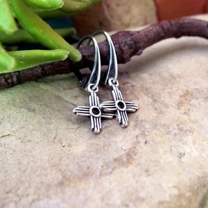 May include: A pair of sterling silver drop earrings featuring a Zia sun symbol design. Each earring has a curved hook wire and a small, oxidized silver cross-shaped charm with four radiating lines on each side and a circular center cutout.