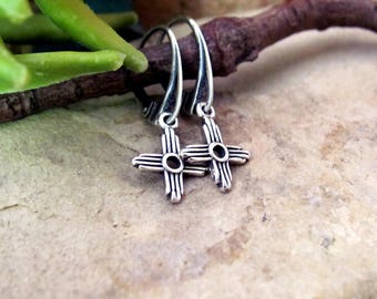 May include: A pair of sterling silver drop earrings featuring a Zia sun symbol design. Each earring has a curved hook wire and a small, oxidized silver cross-shaped charm with four radiating lines on each side and a circular center cutout.