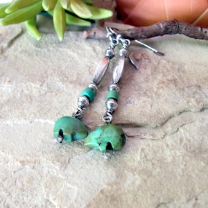 May include: A pair of handcrafted earrings featuring turquoise bear-shaped pendants. The earrings have silver-toned metal accents, including elongated beads and small round beads, and turquoise-colored beads. The earrings are displayed on a textured, light-colored surface.