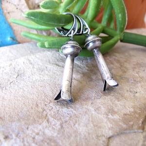 May include: A pair of silver dangle earrings with a rounded top and a pointed bottom. The earrings are hanging from a green plant.