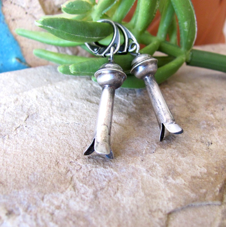 May include: A pair of silver dangle earrings with a rounded top and a pointed bottom. The earrings are hanging from a green plant.