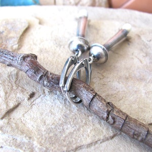May include: Silver earrings with a unique design featuring a long, cylindrical drop with a rounded top and a smaller, round ball at the bottom. The earrings are connected to a silver hook.