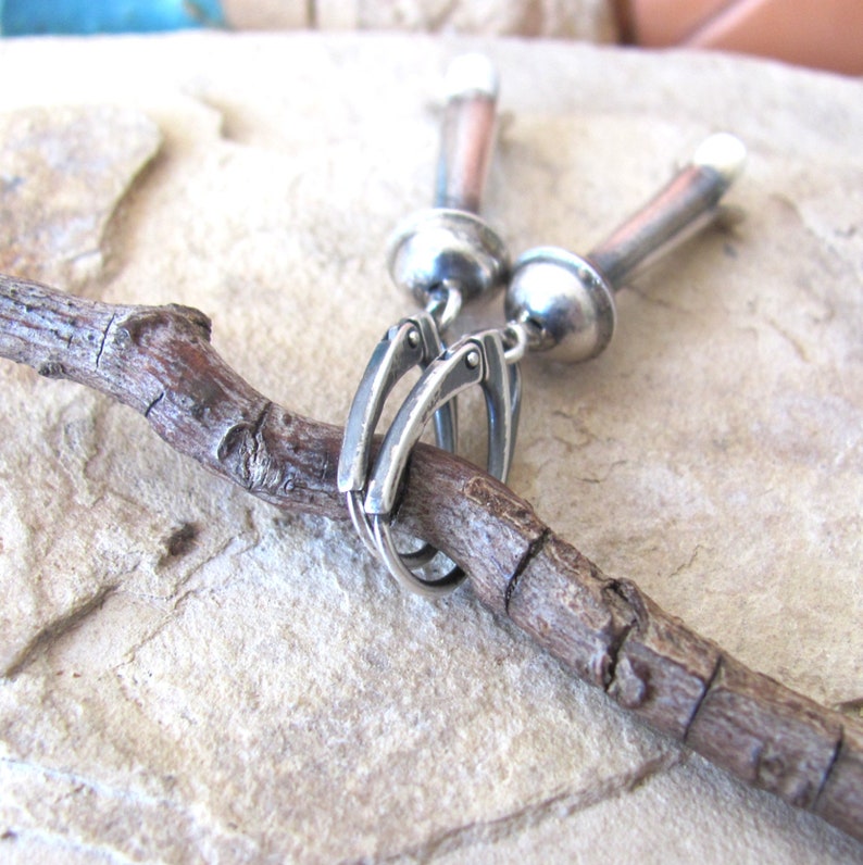 May include: Silver earrings with a unique design featuring a long, cylindrical drop with a rounded top and a smaller, round ball at the bottom. The earrings are connected to a silver hook.