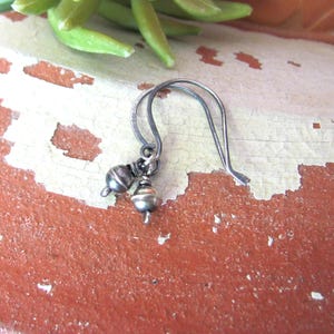 May include: A single earring featuring a dark silver-toned metal hook wire and two small, round, metallic beads suspended from the bottom. The beads have a smooth, polished finish with a subtle sheen, and the metal components exhibit a darkened, oxidized appearance throughout the entire piece.