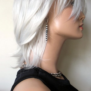 May include: A silver beaded dangle earring with a silver hook. The earring is worn by a person with white hair.