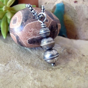 May include: A silver necklace with three stacked, round, silver beads. Each bead has a unique, intricate design. The necklace is on a chain and is resting on a brown and black patterned rock.