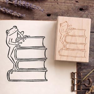 May include: A wooden stamp featuring a frog climbing a stack of books. The design is also printed in black ink on a piece of paper. The stamp and drawing are set against a rustic wooden background with lavender sprigs.