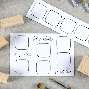 May include: A collection of art supplies including blank color swatch cards, wooden stamps with text, and paint tubes. The cards have square outlines for color swatches, with the phrases "les couleurs", "my colors", and "swatches" printed on them.