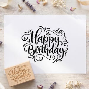 May include: A rectangular wooden rubber stamp featuring the text Happy Birthday in a decorative script font, positioned next to a white card displaying the same Happy Birthday text in black ink, surrounded by sprigs of lavender and baby's breath.