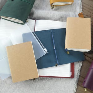 May include: An assortment of leather-bound journals and notebooks in various colors, including green, brown, and purple. Some are open, revealing lined pages, while others are closed with elastic closures. Clear plastic sleeves and a blue pen are also visible.