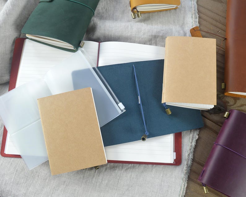 May include: An assortment of leather-bound journals and notebooks in various colors, including green, brown, and purple. Some are open, revealing lined pages, while others are closed with elastic closures. Clear plastic sleeves and a blue pen are also visible.