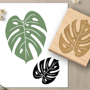 May include: A wooden stamp with a carved monstera leaf design, alongside green and black leaf prints on white paper. The stamp is rectangular and the leaves have a tropical theme.