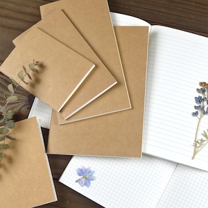 May include: A collection of notebooks with plain brown covers, some stacked and others open to reveal white pages with either horizontal lines or a grid pattern. A sprig of dried eucalyptus and a pressed blue flower are placed on the pages.