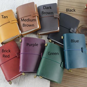 May include: A collection of leather journals in various colors, including tan, medium brown, dark brown, black, brick red, purple, green, and blue. Each journal has a strap closure and a brass-colored metal accent.