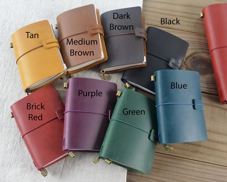 May include: A collection of leather journals in various colors, including tan, medium brown, dark brown, black, brick red, purple, green, and blue. Each journal has a strap closure and a brass-colored metal accent.