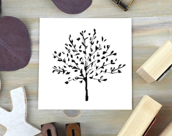 Könnte beinhalten: Ein schwarz-weißer Baumstempel auf einem weißen Quadrat. Das Baumdesign zeigt einen Stamm und Äste mit Blättern. Der Stempel ist von Holzblöcken und einem lila Blatt auf einer hellen Holzoberfläche umgeben. Der Stempel ist für Bastelarbeiten.