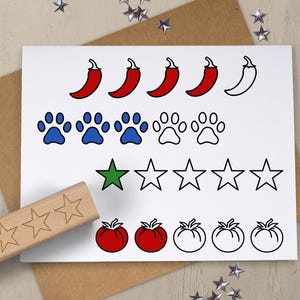 May include: A white card with illustrations of red chili peppers, blue and white paw prints, green and white stars, and red tomatoes. A wooden stamp with star shapes is in the foreground. The card is on a brown envelope.