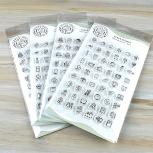 May include: Four clear stamp sets with various planner icons, including a book, clock, and shopping cart. Each set is packaged in a clear plastic sleeve. The text "CLEAR STAMPS Planner Icons - Set 1" is visible on the packaging.